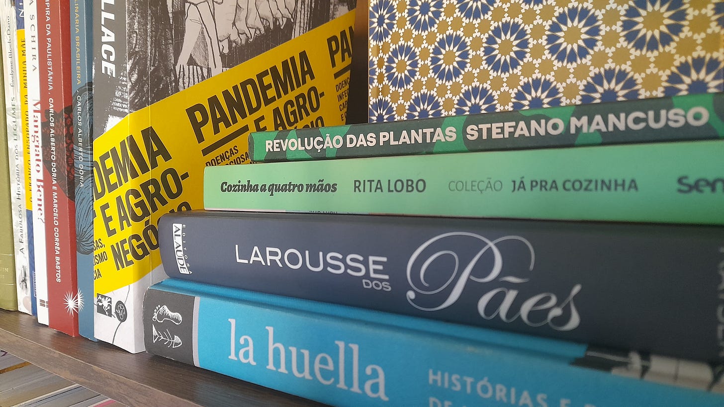 Livros Revolução das plantas, Cozinha a quatro mãos, Larousse dos pães e La Huella sobrepostos em uma prateleira de uma estante marrom. Ao fundo, o livro Pandemia e agronegócio, seguido por outros títulos sobre comida que estão fora de foco e ilegíveis. Livros Revolução das plantas, Cozinha a quatro mãos, Larousse dos pães e La Huella sobrepostos em uma prateleira de uma estante marrom. Ao fundo, o livro Pandemia e agronegócio, seguido por outros títulos sobre comida que estão fora de foco e ilegíveis.