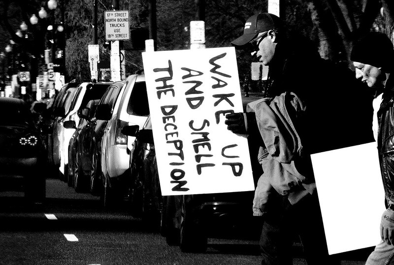 A black and white photo of a protest. A man holds a sign sideways, which reads in blobby letters, "WAKE UP AND SMELL THE DECEPTION"