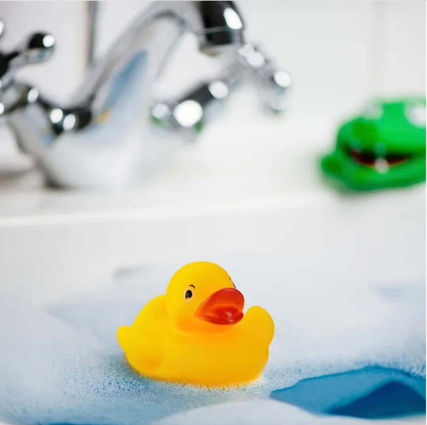 Una papera di gomma gialla che galleggia in una vasca da bagno piena di schiuma blu. Sullo sfondo, un rubinetto cromato sfocato e un dinosauro giocattolo verde. Una papera di gomma gialla che galleggia in una vasca da bagno piena di schiuma blu. Sullo sfondo, un rubinetto cromato sfocato e un dinosauro giocattolo verde.