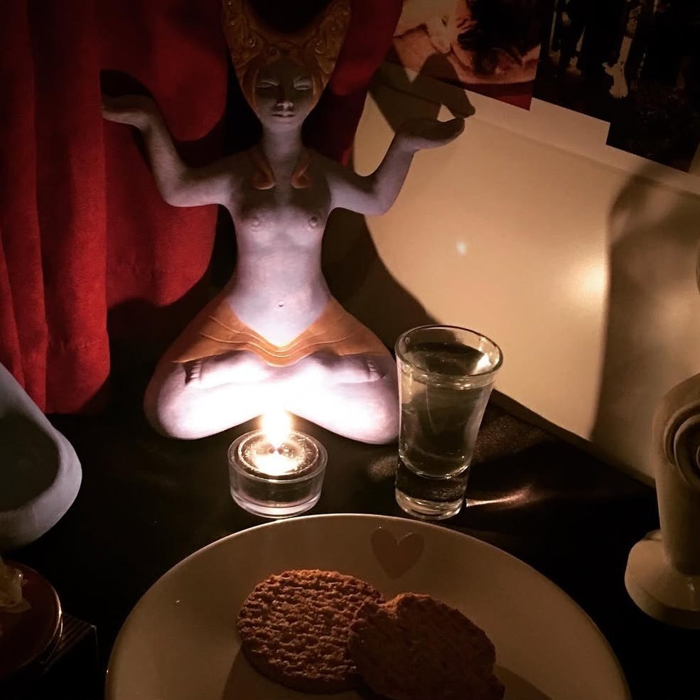 A single lit tea light candle casts lights and shadows on an altar to the ancestors. Behind the candle is a statue of a seated Goddess with arms outstretched to the sides, palms upright. To the right of the candle is a clear shot glass filled with water. Before the candle is white plate with two oat biscuits on it. To the wall behind the altar is a photo collage of animals and people: some of the author’s Beloved Dead, who are being honoured at this time.