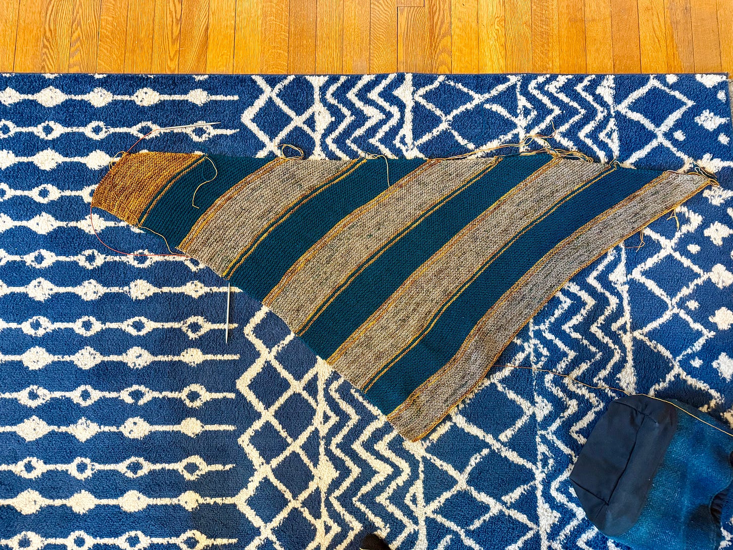 Triangle shawl with teal and grey stripes, and gold accents, laying flat on a blue and white patterned rug.