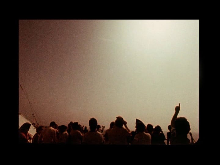 The second of four shots from a sequence in Peter Rose's The Man Who Could Not See Far Enough depicting a crowd on a ship watching a solar eclipse. The sky has started to faintly darken; one onlooker points up toward the sun.