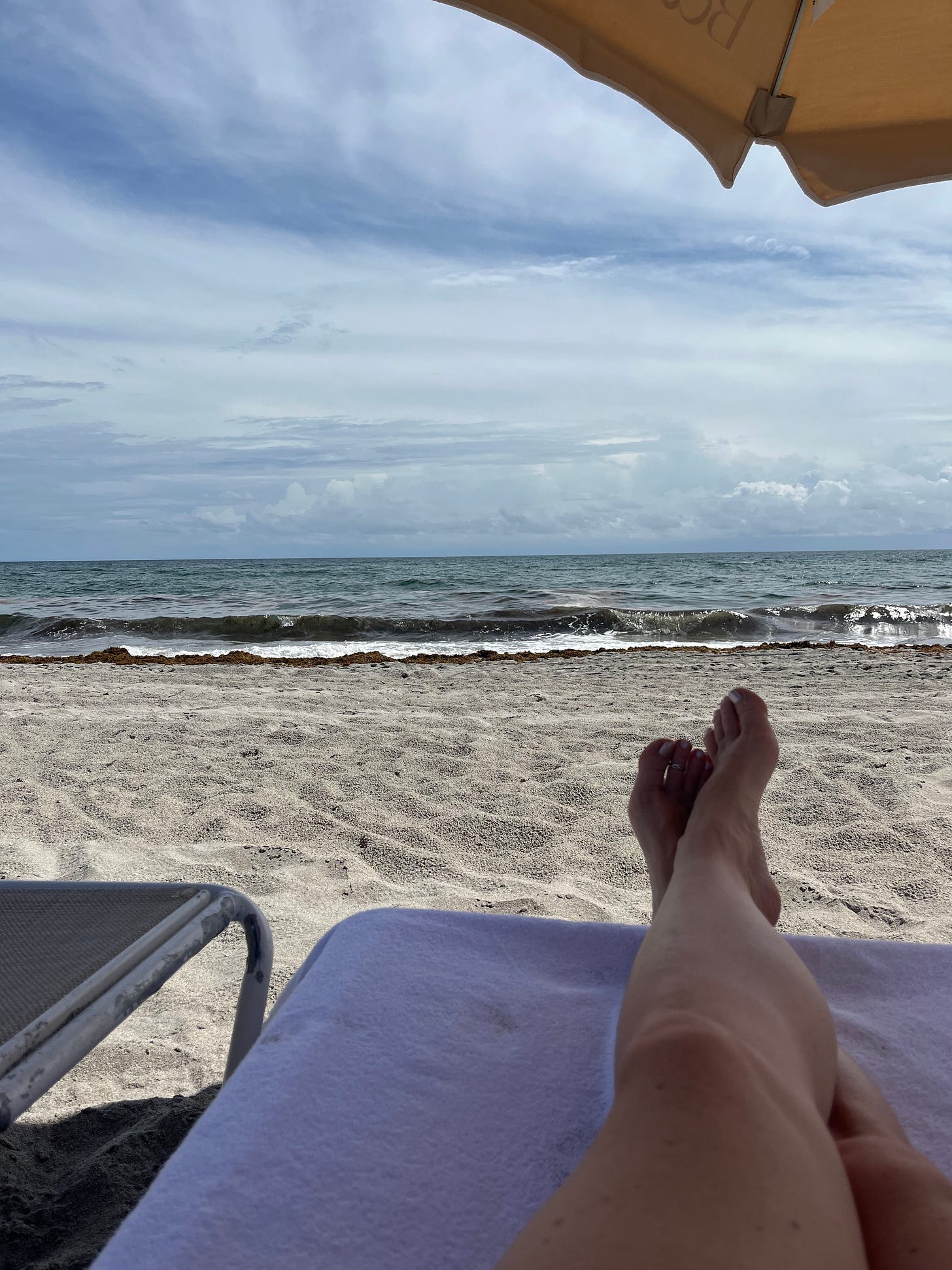 a white capped wave rolls into the beach below a sky strewn with thin clouds; the author's legs are resting in the foreground on a beach chair, and a small portion of a yellow beach umbrella is seen in the upper right quadrant
