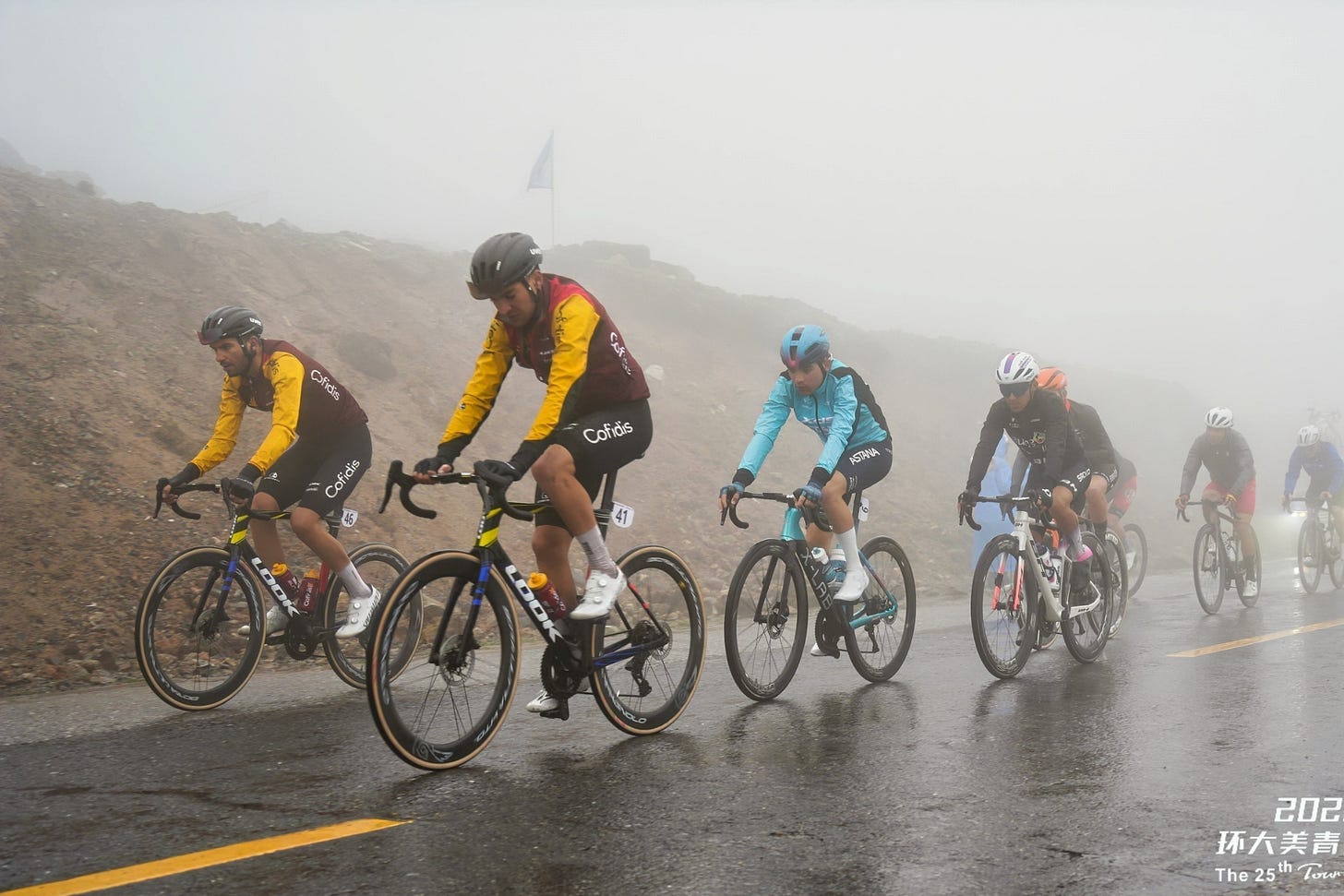 The peloton, wearing rain jackets, plows through fog and rain with their heads down.