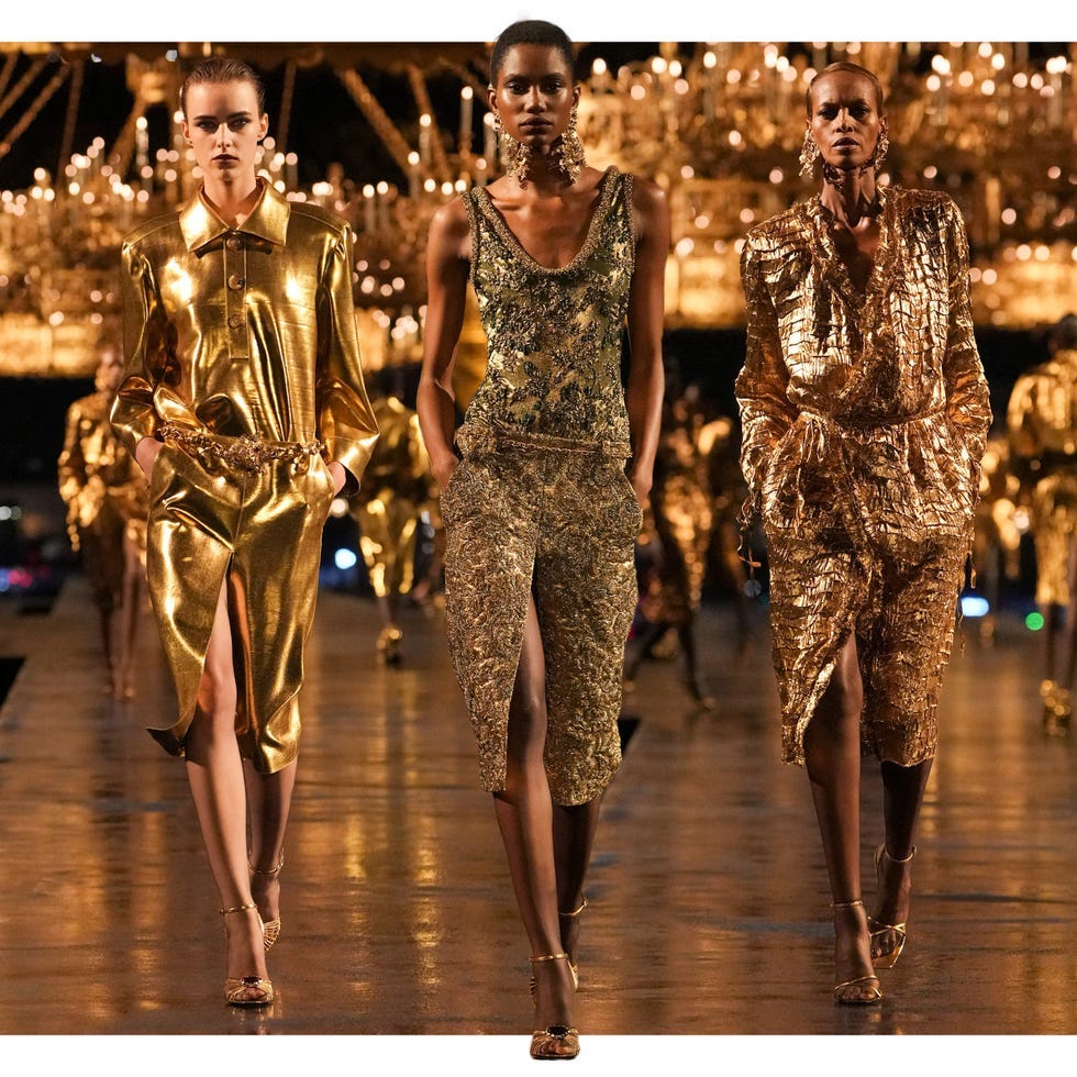 Three models showcase glamorous golden outfits on a runway illuminated by chandeliers.