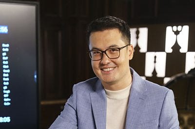 David Howell wearing a grey blazer smiling at the camera. The background is blurred