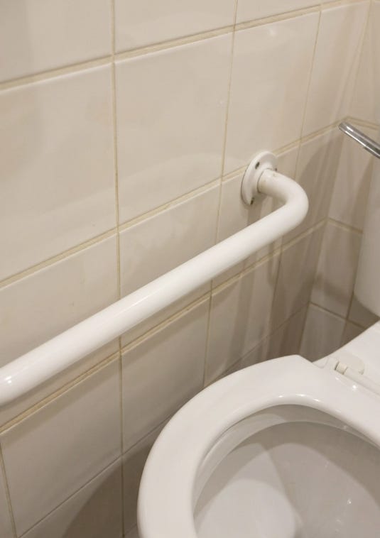 Accessible bathroom in car showroom, white tiled walls, a white grab rail and a toilet positioned very close to the wall just under the grab rail. The flush handle almost touched the wall too.