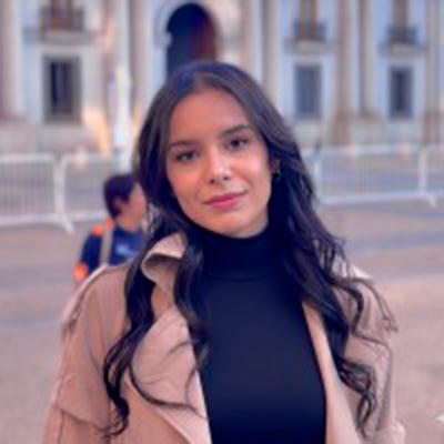 Mulher branca de cabelos pretos em compridos e um sobretudo cáqui em uma praça. Sorri levemente para a câmera Mulher branca de cabelos pretos em compridos e um sobretudo cáqui em uma praça. Sorri levemente para a câmera