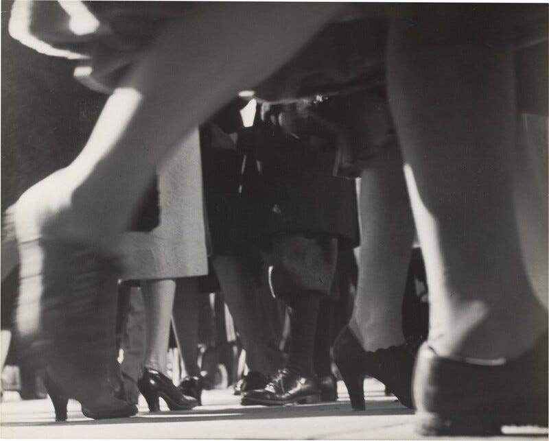 This is a photograph of people walking, captured from a ground-level perspective focusing on their legs and feet. The image depicts a dynamic scene where the motion is evident in the blur of some legs and feet. The photograph captures a variety of footwear, predominantly high-heeled shoes. The composition utilizes low-angle and close-up views. The contrast between the dark and light areas enhances the sense of depth and movement within the image.
