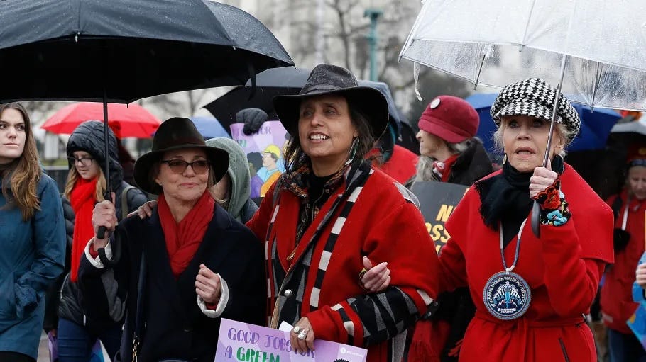 A photo of Winona flanked by Sally Field and Jane Fonda, walking arm in arm at a rally. Winona is wearing a black hat and a red blanket shawl and smiling while holding a sign down by her waist. Sally is holding an umbrella and wearing a black wide brim hat and a black jacket with a red scarf and sunglasses. Jane is wearing a red jacket with a black scarf. She is holding a clear umbrella and wearing a black and white striped hat.