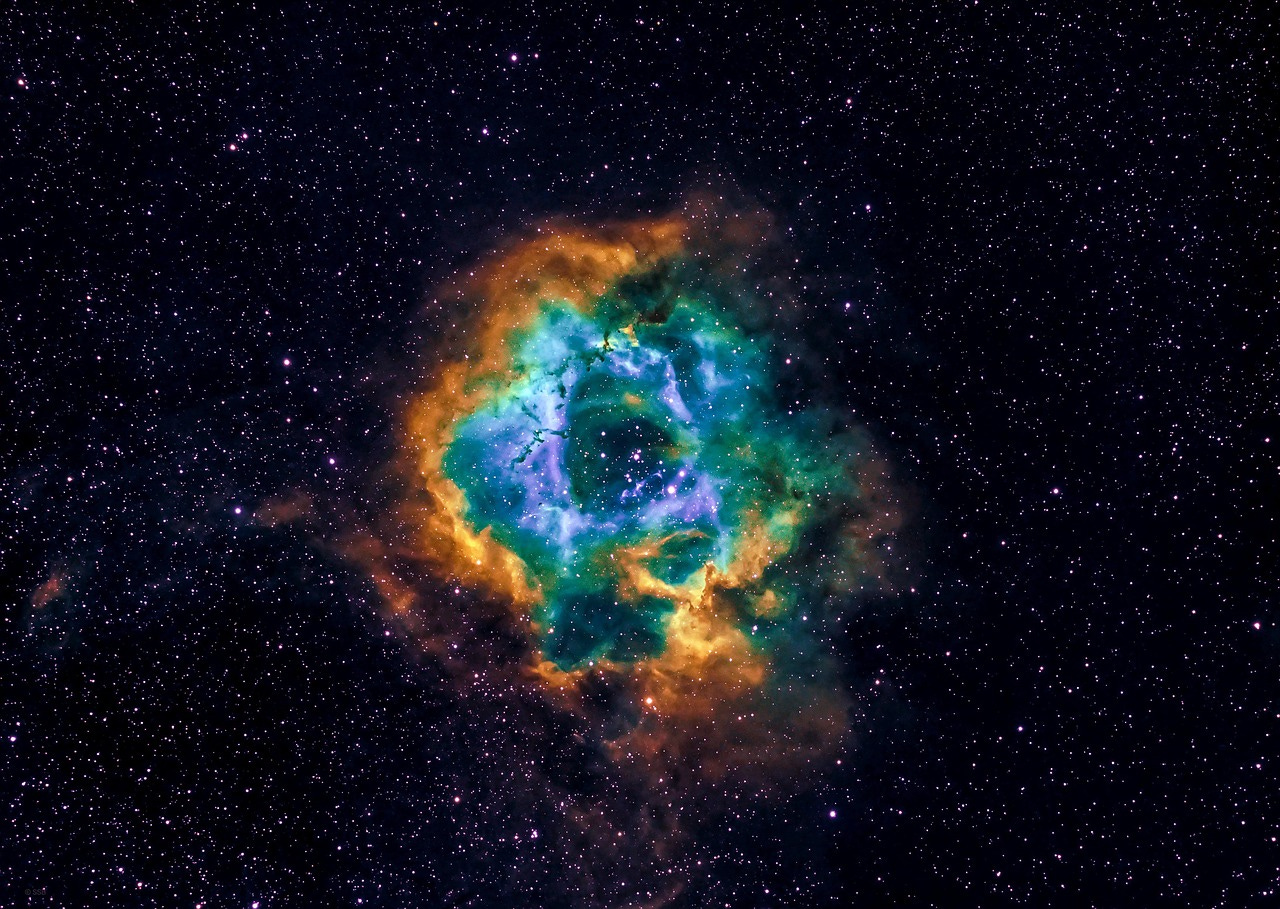 In the center of a star-encrusted expanse of black space sits a colorful nebular of orange, green, and blue in a circular shape. The nebula glows with radiant light.