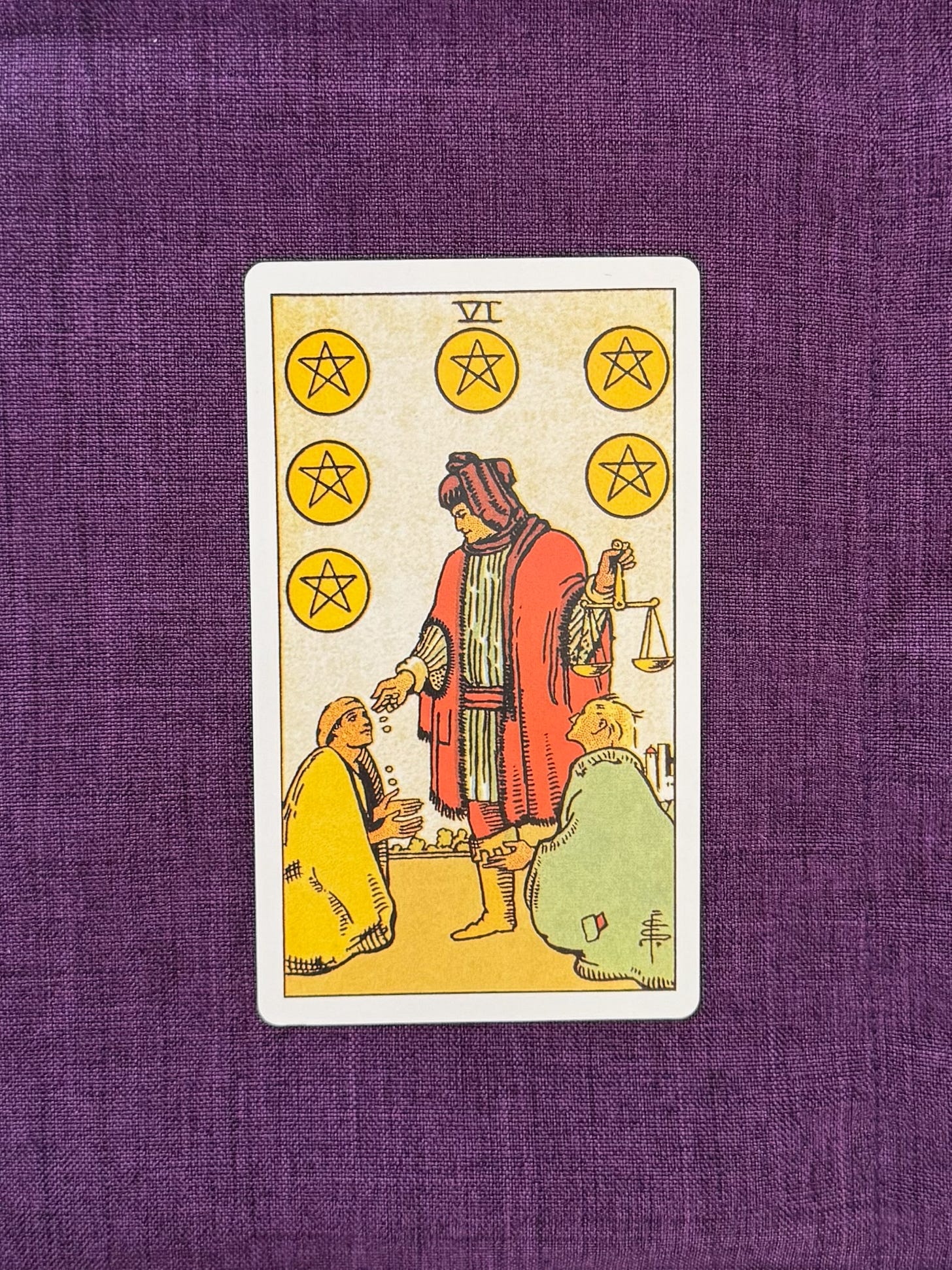 The Six of Pentacles shows a wealthy man dressed in a red robe, handing out coins with his right hand to two beggars who kneel at his feet. In his left hand, he holds a balanced scale.
