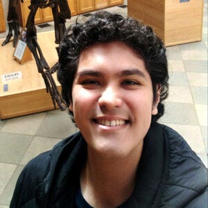 Homem jovem, cabelos volumosos e escuros, usa casaco preto e está à frente de um esqueleto de dinossauro, no Museu de Oxford, Reino Unido Homem jovem, cabelos volumosos e escuros, usa casaco preto e está à frente de um esqueleto de dinossauro, no Museu de Oxford, Reino Unido