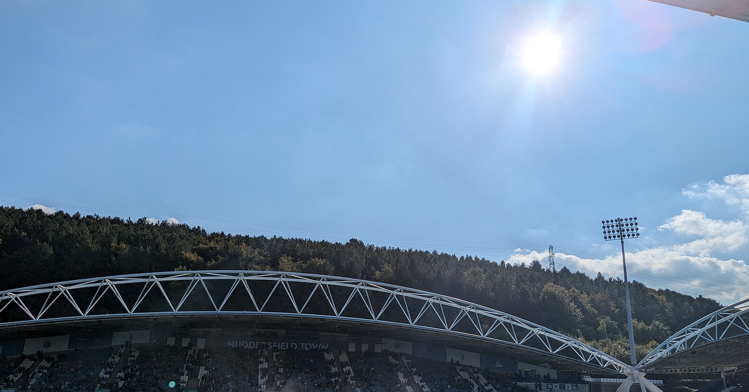 A photograph of Huddersfield Town’s Accu Stadium. The image depicts the top third of the Kilner Bank Stand, with trees behind it, and a bright blue sky. A photograph of Huddersfield Town’s Accu Stadium. The image depicts the top third of the Kilner Bank Stand, with trees behind it, and a bright blue sky.