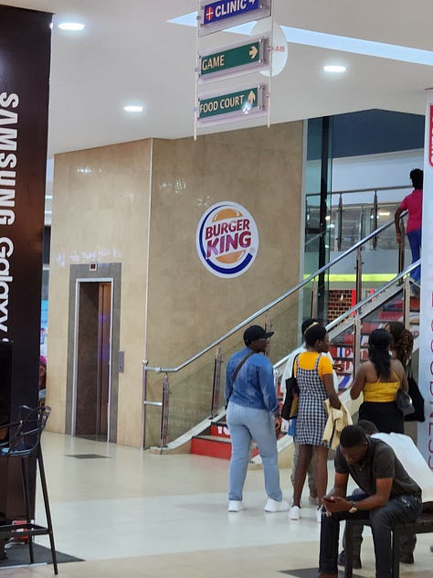Two multicolored high rise apartment buildings, a Burger King sign near a stairwell in the mall, and a table full of fresh plantains on a roadside vendors table.