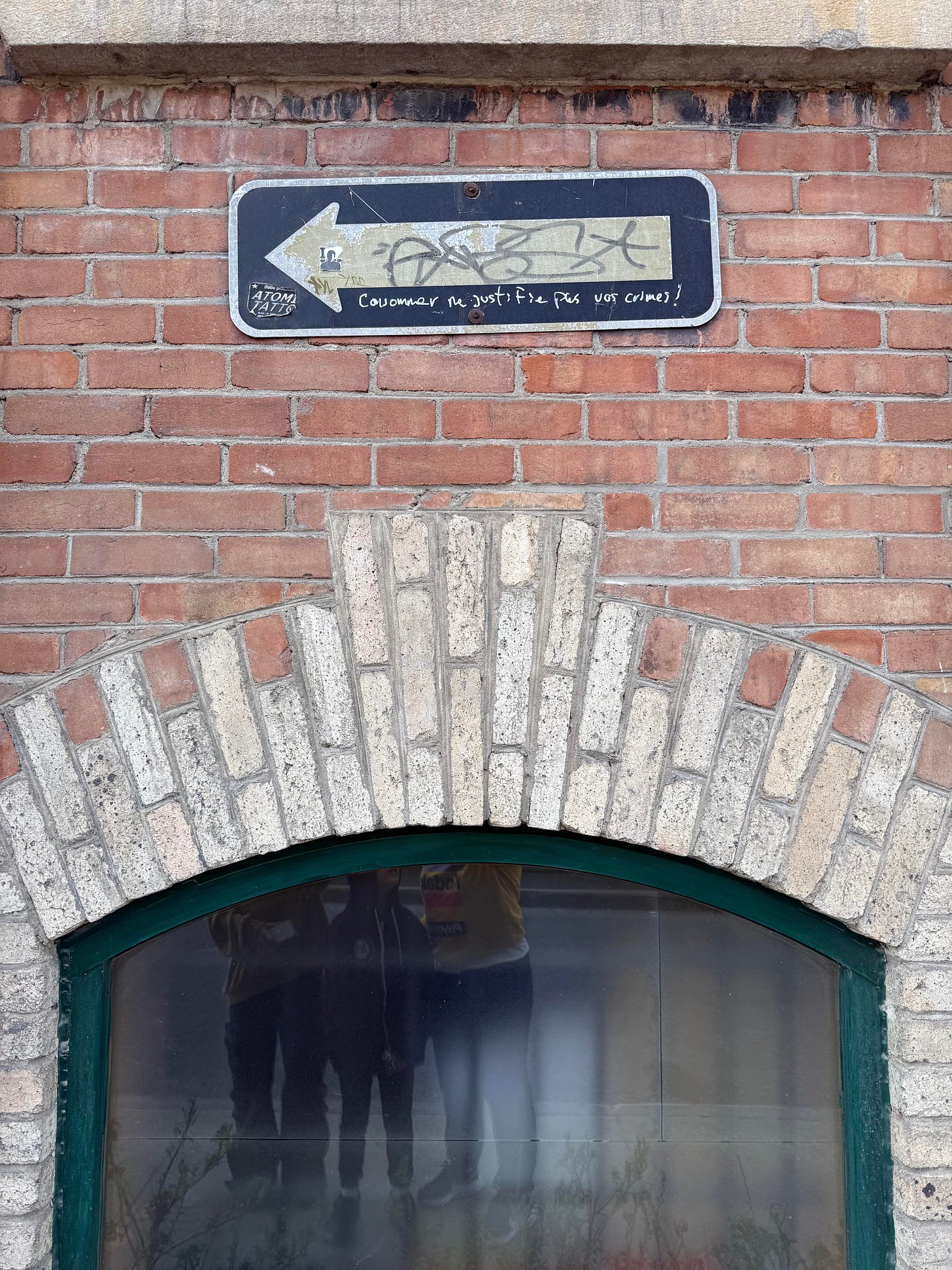 A black arrow sign mounted on a red brick wall above an arched window reads "Consommer ne justifie pas vos crimes!" with graffiti tags overlaid. Three figures are partially visible reflected in the window below.