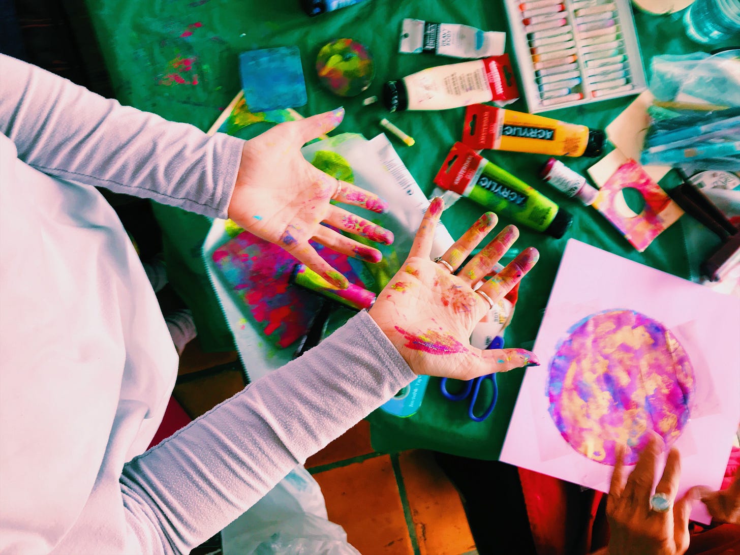 a person in a grey shirt holding out their paint covered hands over a table covered in art supplies and round paintings. 