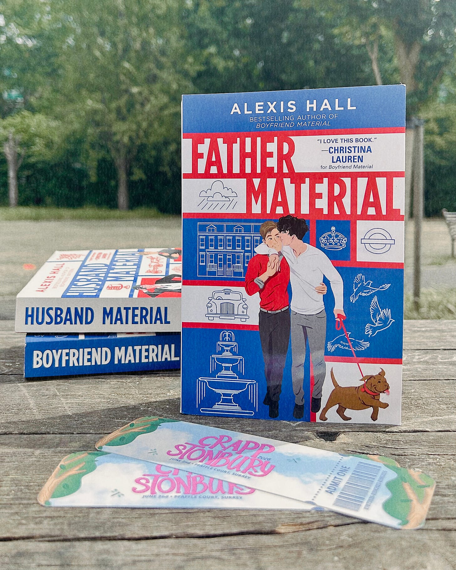 There’s a wooden picnic table with a paperback copy of Father Material by Alexis Hall standing upright with the cover facing outwards. Behind Father Material are paperback copies of Boyfriend Material and Husband Material, both by Alexis Hall, stacked on top of each other with their spines out. In front of Father Material are two CRAPPstonbury bookmarks.