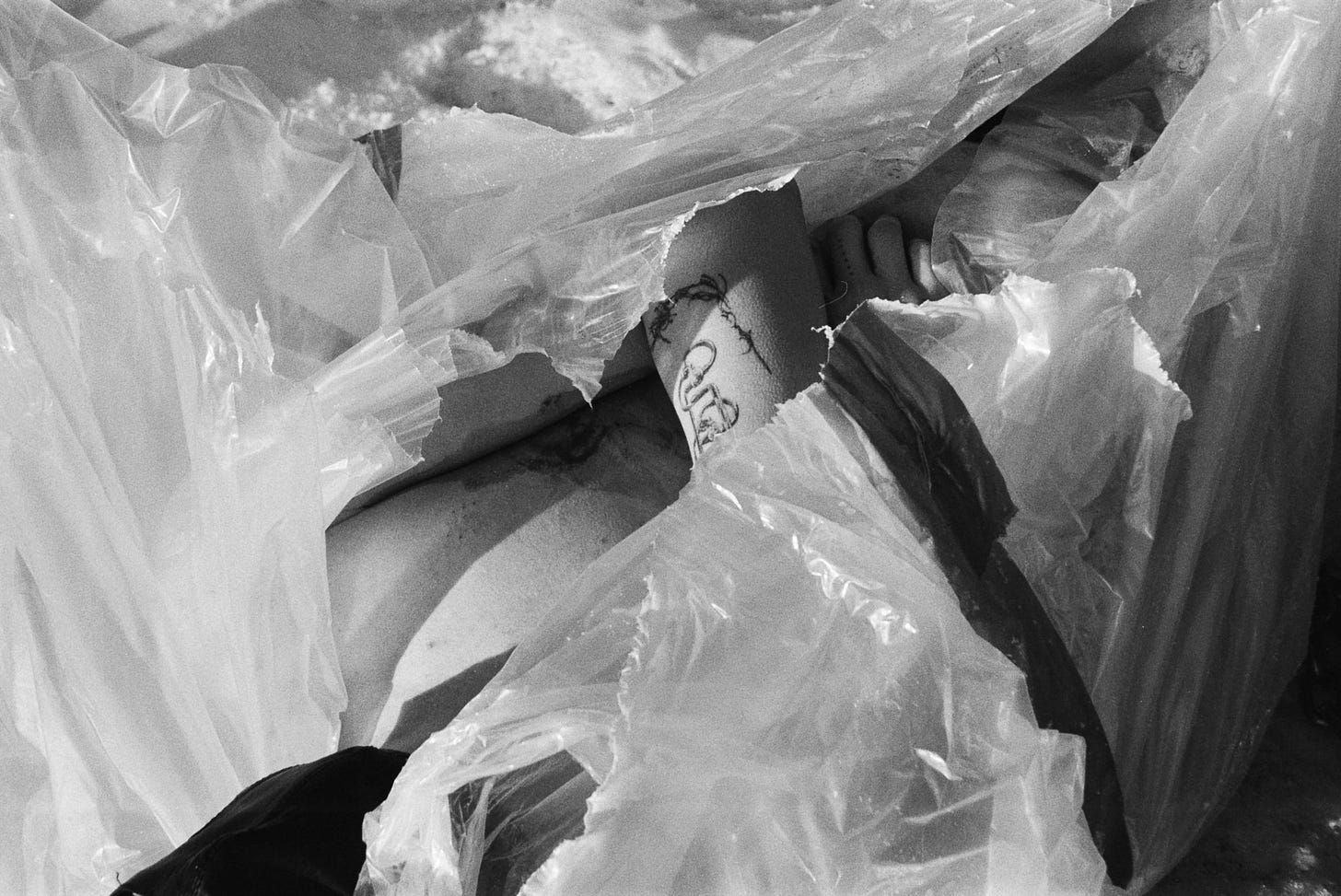 A pale body with tattoos in plastic wrap with snow on the ground. A gloved hand reaches towards the plastic wrap to rip it open further.
