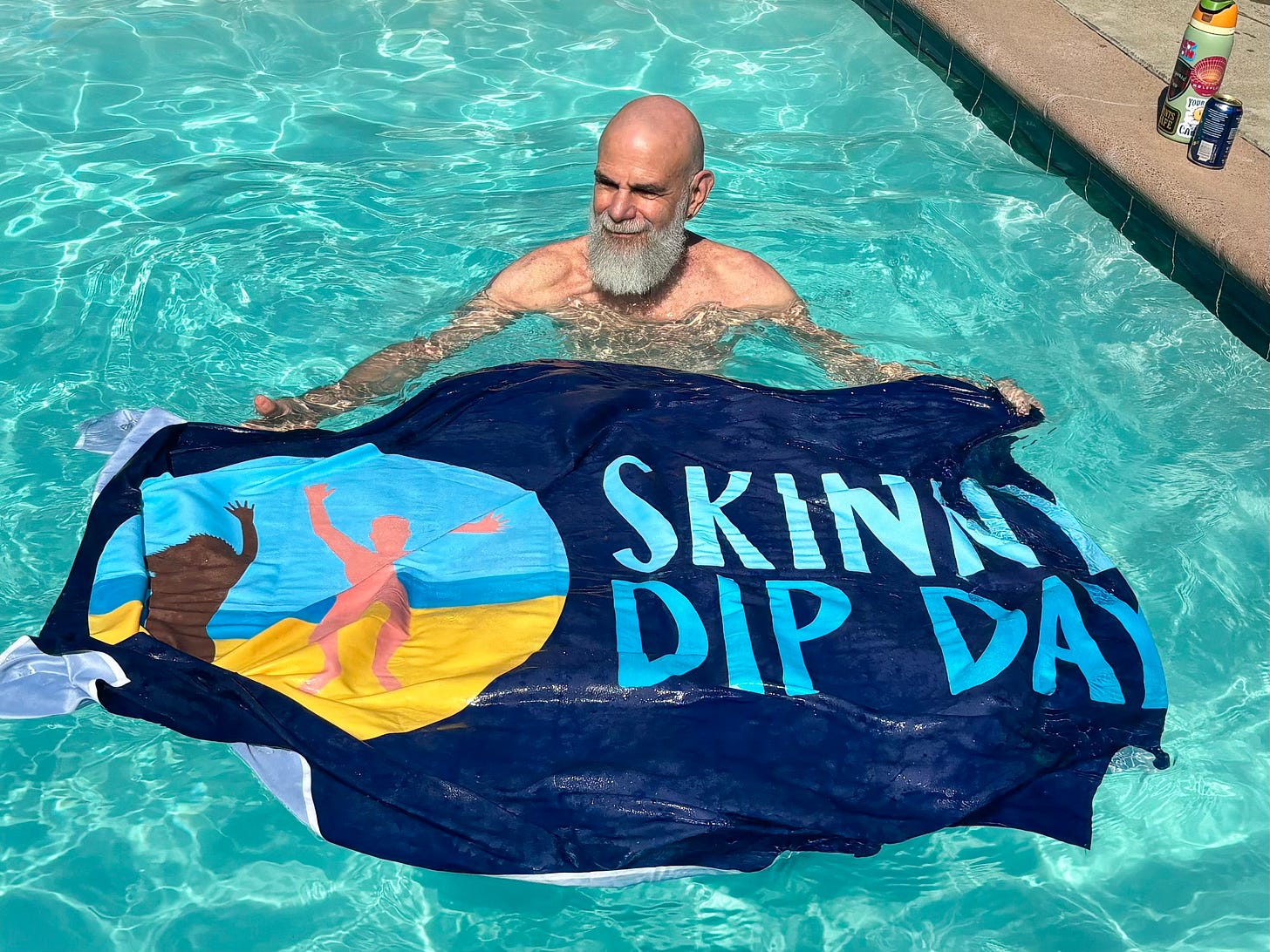 Andy Tabbat floats in a swimming pool holding a Skinny Dip Day flag partially submerged in the water. Andy Tabbat floats in a swimming pool holding a Skinny Dip Day flag partially submerged in the water.