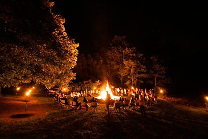 A huge bonfire at night with many inhabited chairs placed in a circle around the fire. There are a few people standing up around the fire. There are several trees. It is night time A huge bonfire at night with many inhabited chairs placed in a circle around the fire. There are a few people standing up around the fire. There are several trees. It is night time