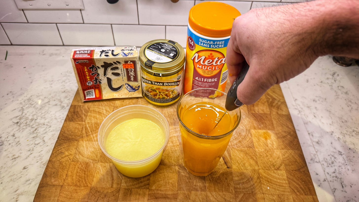 Two rows of items. In the back, packages of instant dashi, Better than Bouillon chicken stock concentrate, and Metamucil. In the front there is a deli container of well-blended chicken stock and a glass of Metamucil and water being blended with a milk frother. The liquid has formed a vortex around the frother.