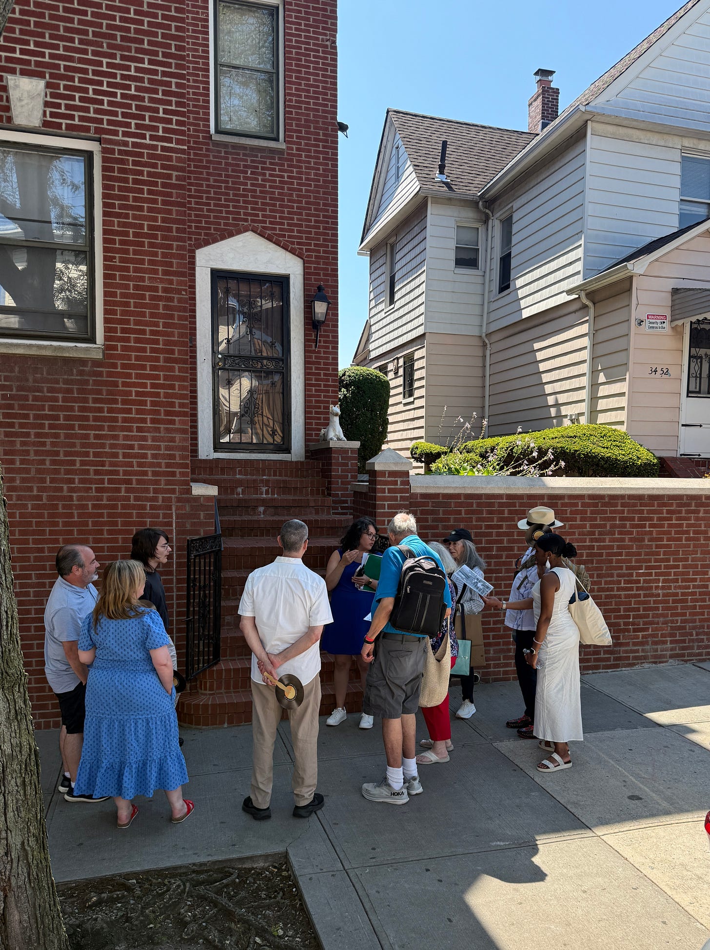 In front of a brick two-story house, a group of people stand in a semicircle on the sidewalk, listening to a genius say genius things.