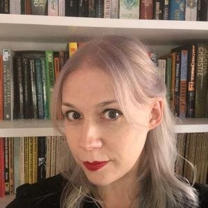 Photo of A K Benedict. White woman with ight hair with fringe. Bookshelf in the background