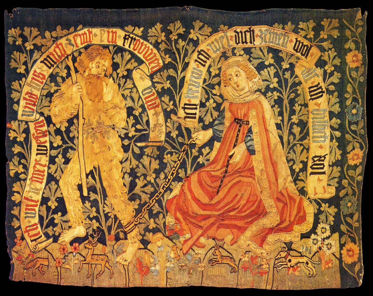 A tapestry showing a bearded man covered in a hair suit, with a woman in a red dress, who is holding a chain connected to the wild man's leg. Scrolls with words surround both figures, and leaves and flowers are the ground for the tapestry