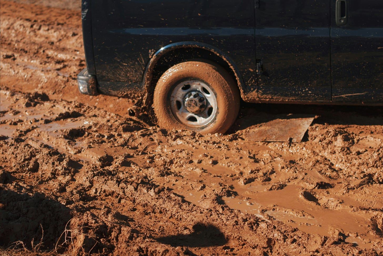 The rear right wheel of a van is centered in the frame, which is mostly red-brown mud, trapping the wheel of the van.
