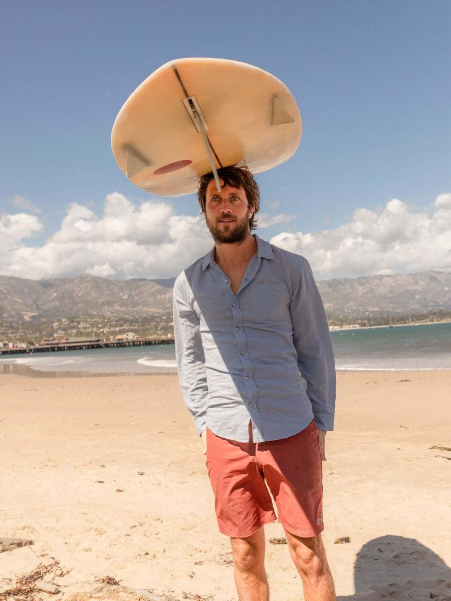 a man balances a surfboard on his head while wearing a blue linen shirt on a beach a man balances a surfboard on his head while wearing a blue linen shirt on a beach