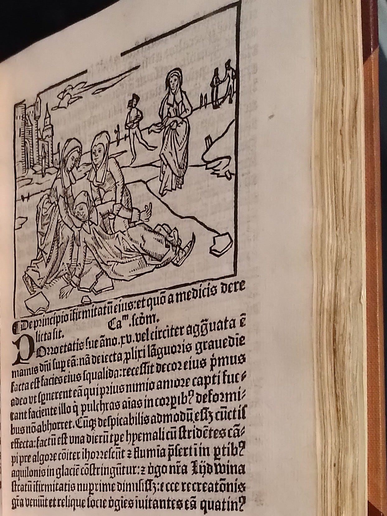 A page of an old Latin printed book with a black and white woodcut: two women kneel on the ice to lift a fallen girl who still has a skate on her foot, while a skater glides past in the background near a town gate. A page of an old Latin printed book with a black and white woodcut: two women kneel on the ice to lift a fallen girl who still has a skate on her foot, while a skater glides past in the background near a town gate.