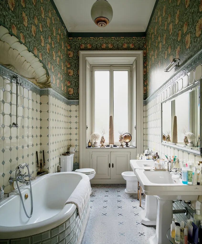 A bathroom with a floral wallpaper at top, and tiled walls at the base. There are two sinks, side by side, a tilet, bidet and built-in bathtub.