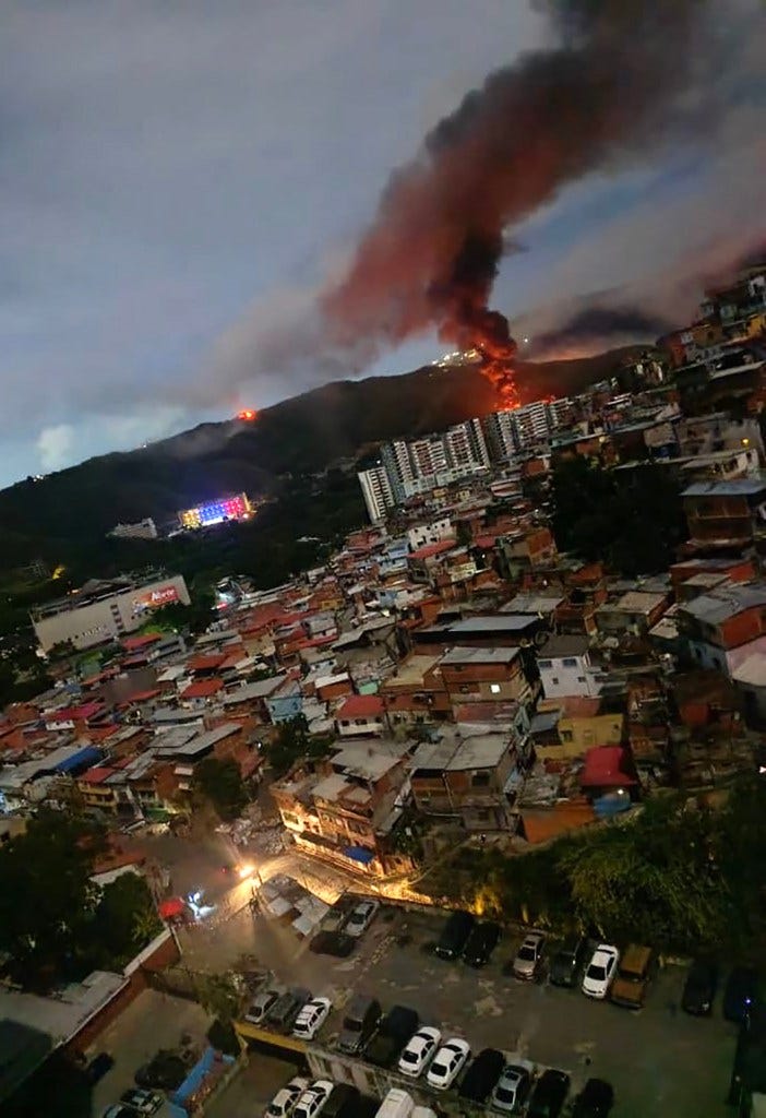 Fire and smoke rising from Caracas mountainside in the distance, with city buildings and homes in the foreground.
