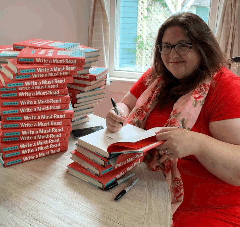 Author AJ Harper signing her book: Write A Must-Read. She's wearing a bright red dress and a a pink scarf and sitting in front of stacks of books.