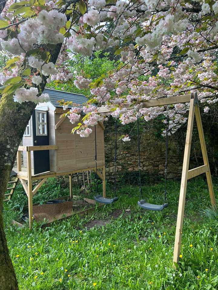 Four photos of a walled French garden in spring: a canopy tent on the grass under blossoming trees, a wooden playhouse and swing set beneath a cherry tree in full pink bloom, a young girl planting seedlings in a raised bed beside a wheelbarrow, and a trampoline framed by climbing roses and a ribbon archway.