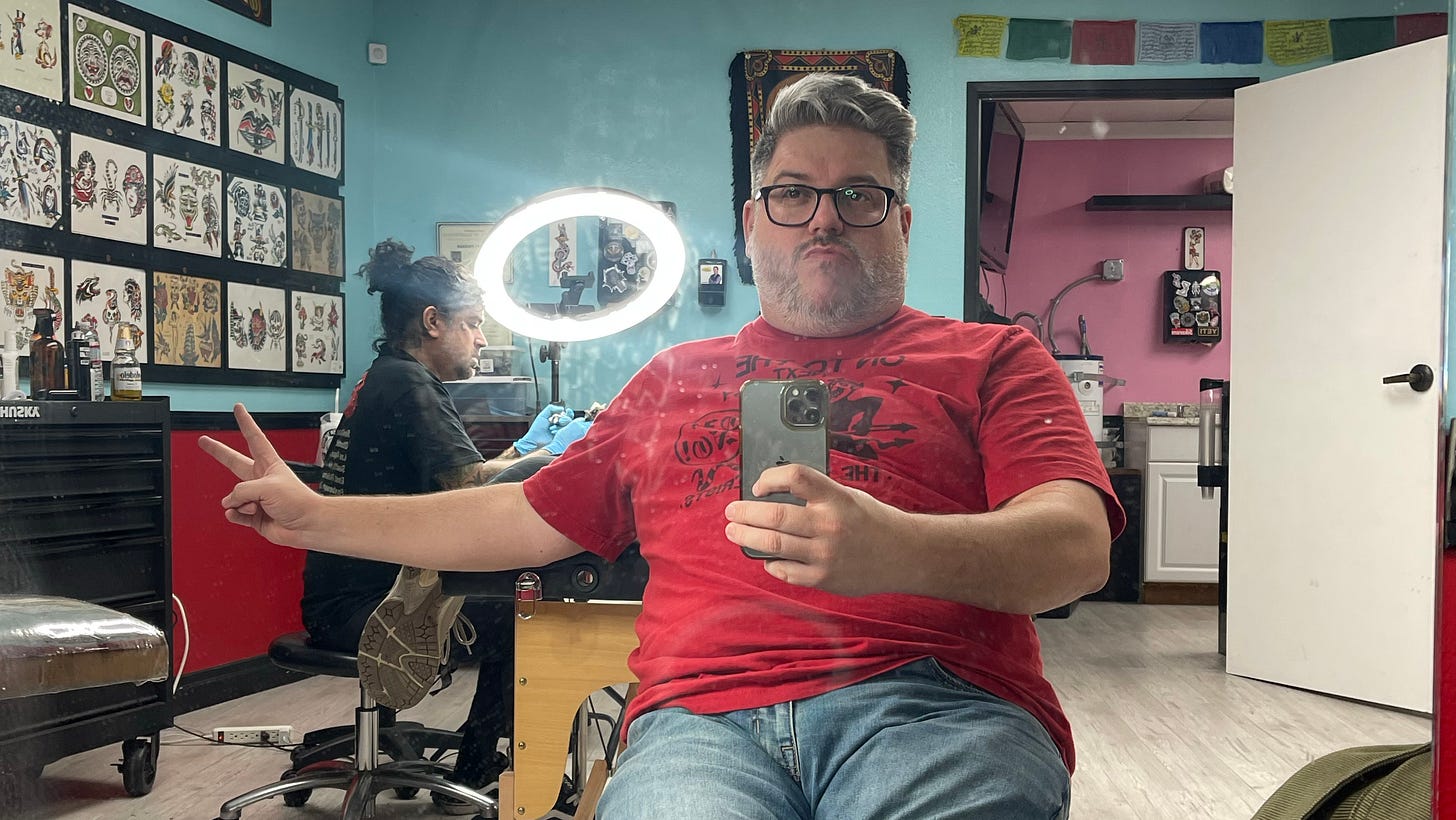 Me hanging out in a tattoo shop, with my tattoo artist friend tattooing in the background. It's another mirror selfie and this time I am chucking the deuce and trying to look cool and failing.