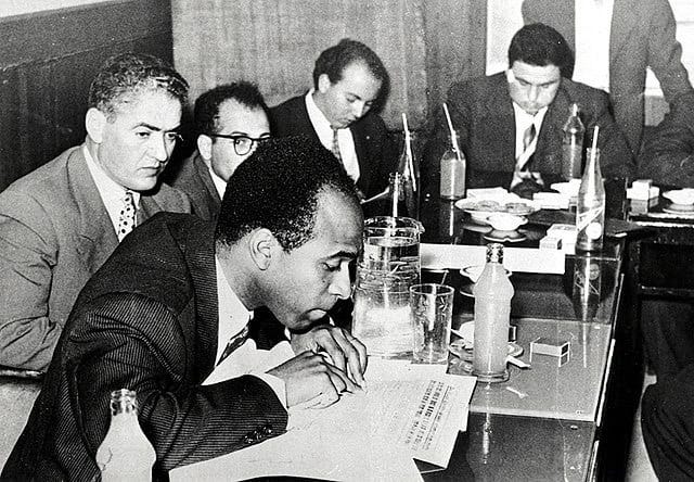 Black-and-white 1959 photo of Frantz Fanon and four other men sitting at a conference table covered with papers and drinks. Fanon is in the foreground reading.