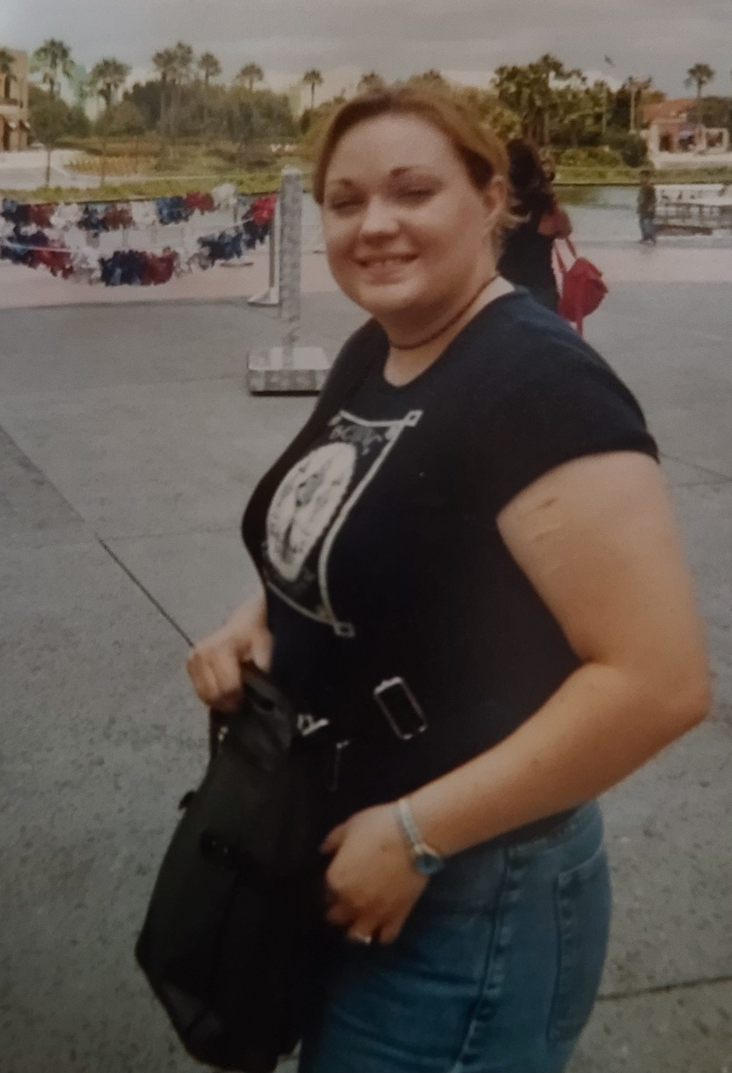 White young woman with her hair pulled back, dark t-shirt and jeans, facing the camera from the side with a little smirk. You can visibly see scars on her upper arm in the foreground from self-harm. She has a messenger bag slung across her torso.