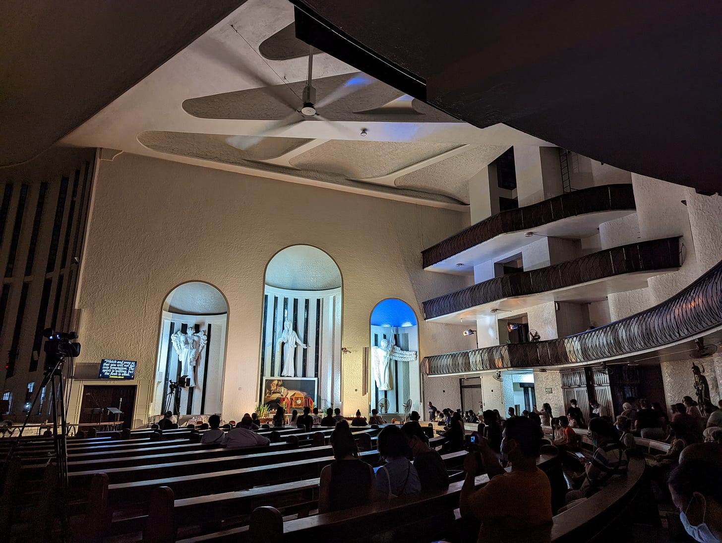 The mostly dark interior of Jesus the Divine Word Church showing the many people visiting against the lit altar of repose