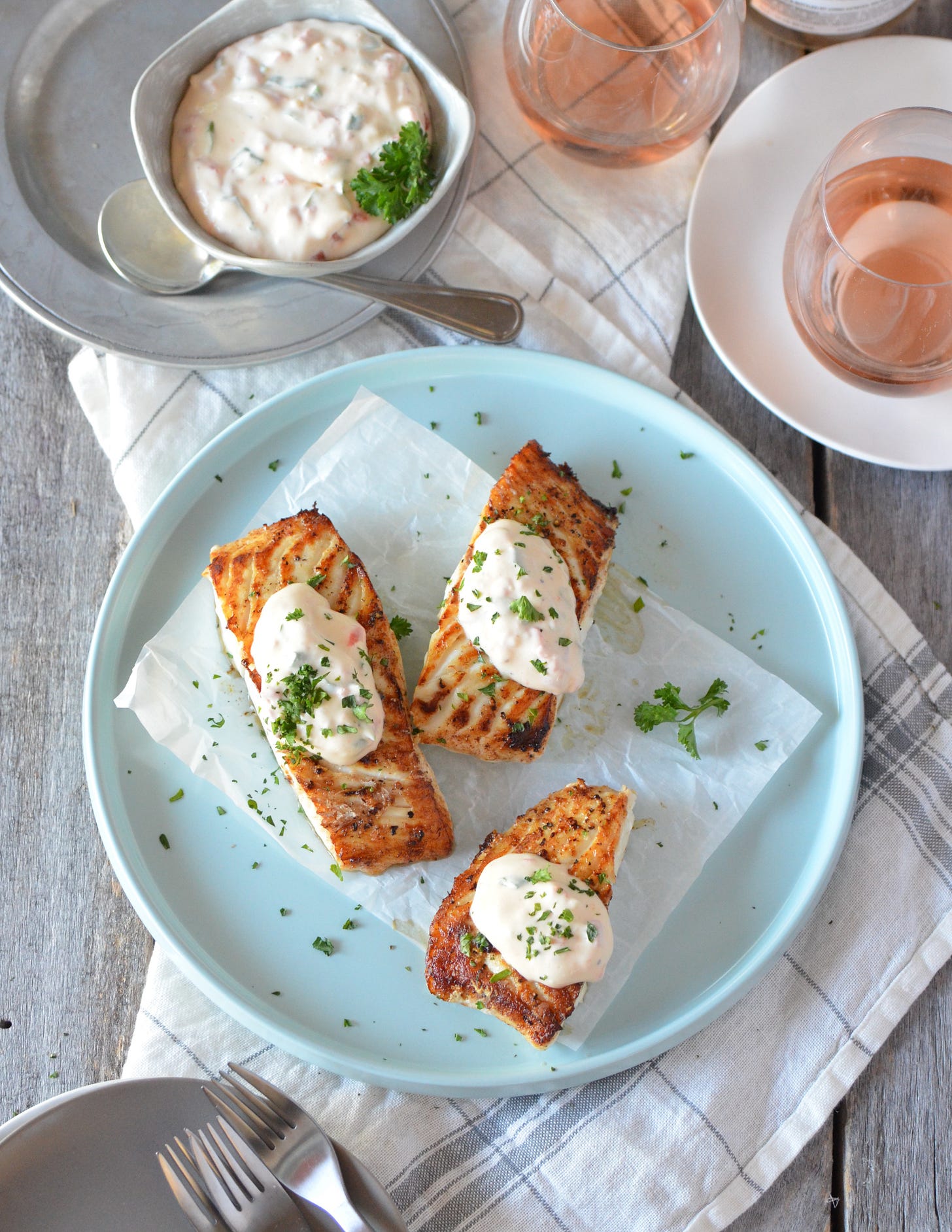 A light blue plate with three pieces of pan-cooked halibut topped with a Whipped Pimento Sauce from the cookbook, For the Love of Seafood by Karista Bennett. The plate is on a gray wood table with a bowl of extra sauce and rose wine in glasses.