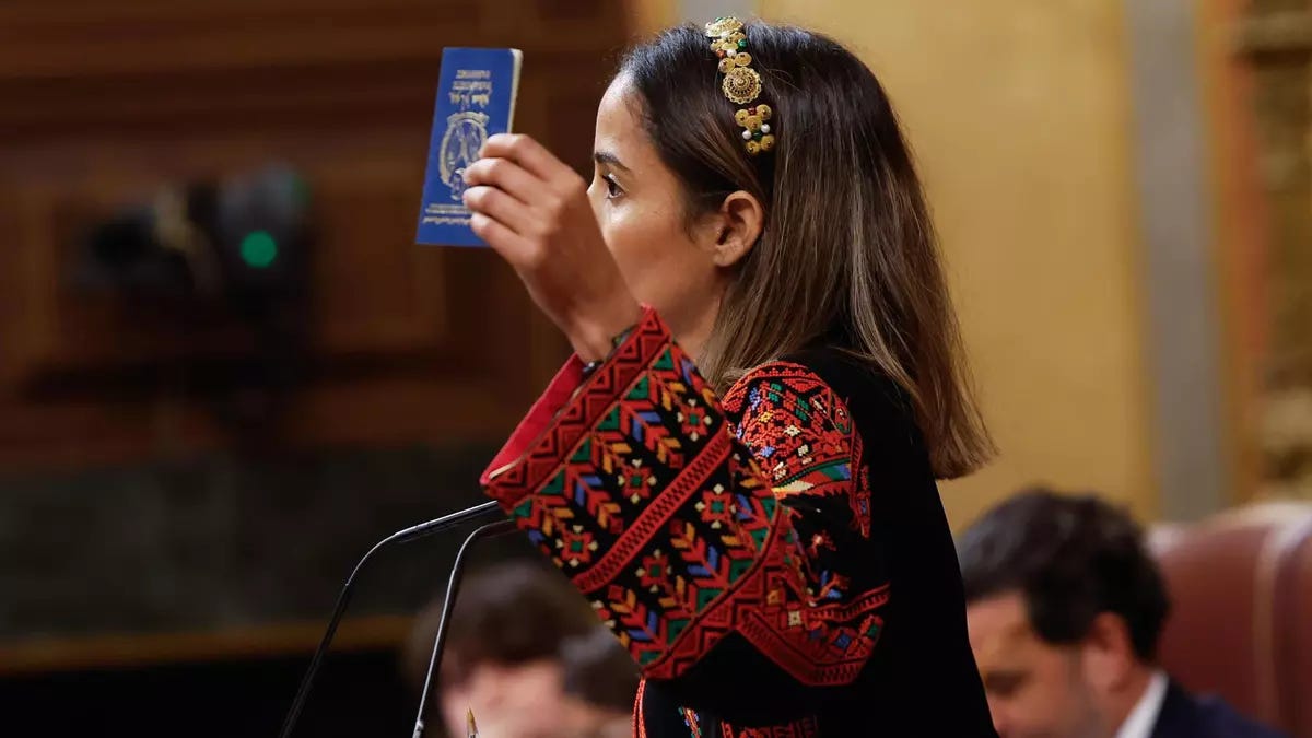 Una donna con lunghi capelli castani, che indossa una fascia dorata e bianca in testa, è vista di profilo mentre tiene in alto un passaporto blu. Indossa un abito nero con ampie maniche decorate con ricami rossi, neri, verdi e gialli: è in un'aula del parlamento, con sedie visibili sullo sfondo e un microfono davanti a lei.