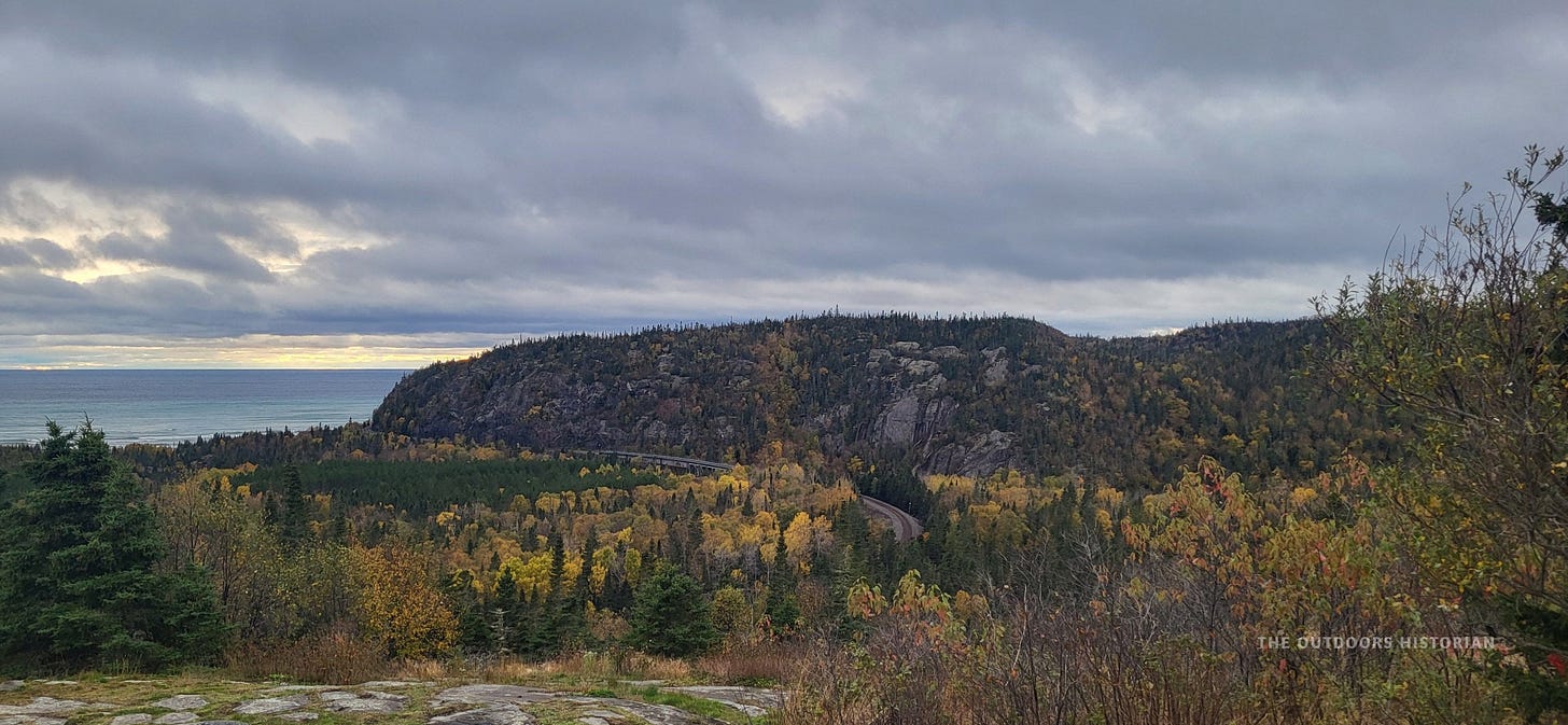 A landscape photo of the Lake Superior shoreline. The sky is filled with heavy, dark grey clouds. A rugged, rocky headland juts out into the lake.