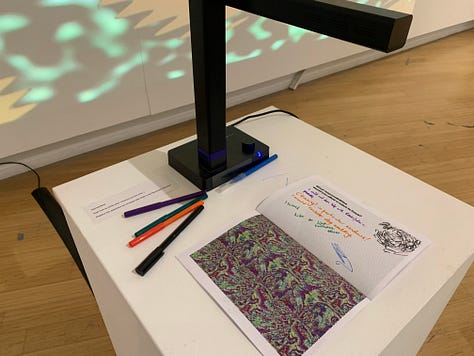 A variety of new media art works using screens, projections, including a large yellow screen and an anaologe cassete with the caption "an actual AI artist" 