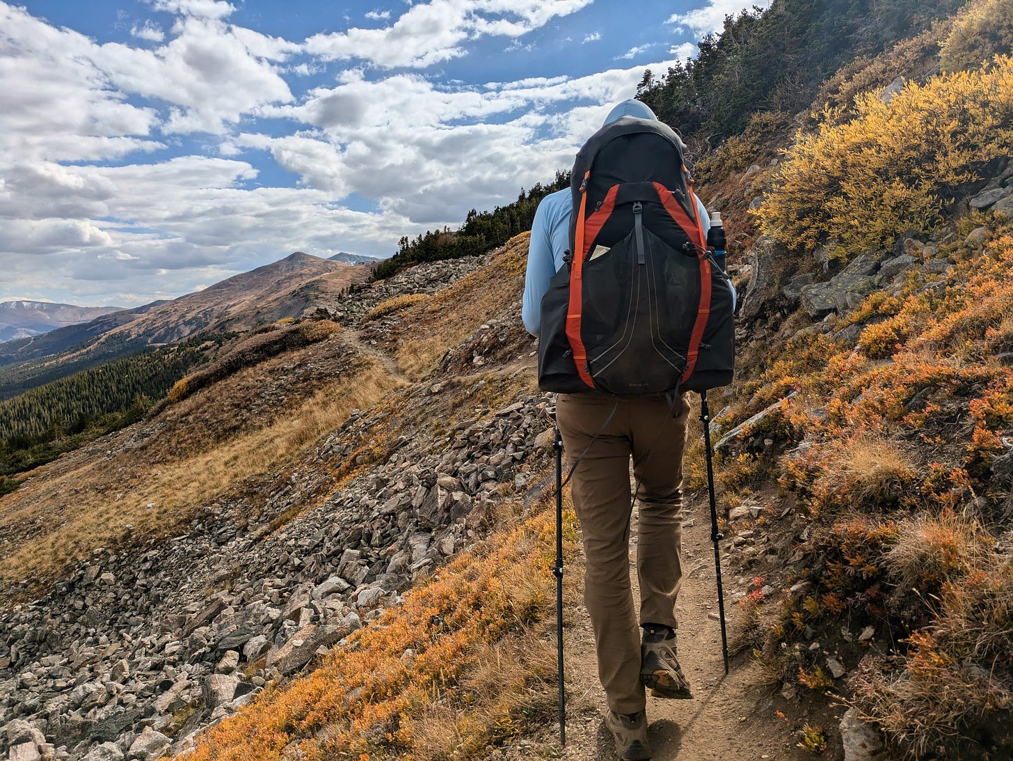 Backpacker hiking along a narrow mountain trail with a full pack and trekking poles, surrounded by rocky terrain and fall foliage in Colorado.