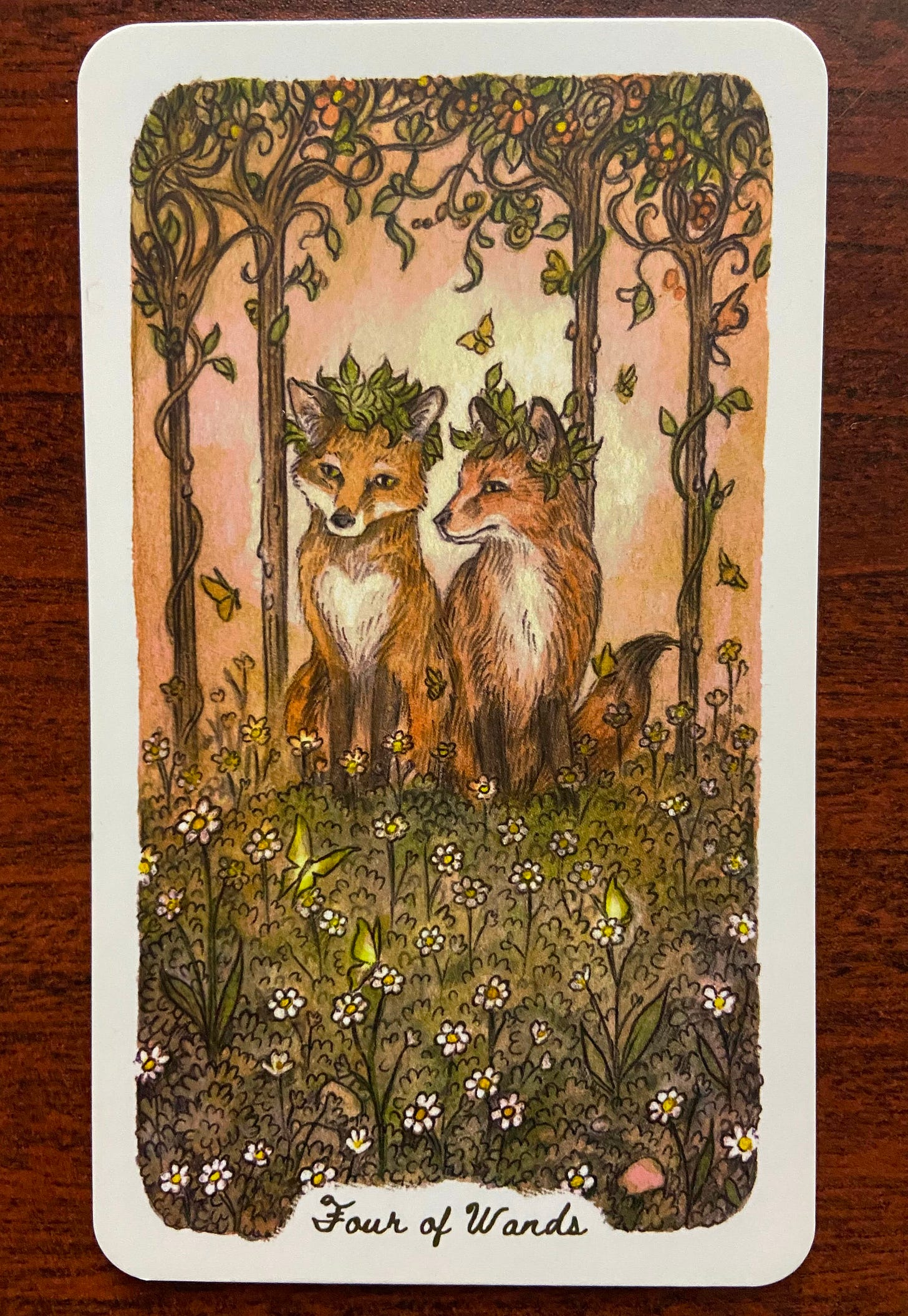 Top card, Four of Wands: Two sweet-faced foxes sit side by side in a field of wildflowers. Their head are crowned by leaves. Behind them stand four slender flowering trees, and a few butterflies are aloft in the air.  Bottom card, Eight of Cups: Under a blazing full moon and a sky of stars, a river flows. A squirrel sits on land facing dark mountains in the distance. Eight “cups”--empty acorns, surrounded by oak leaves–rest in the foreground of the image. The squirrel has left them behind.