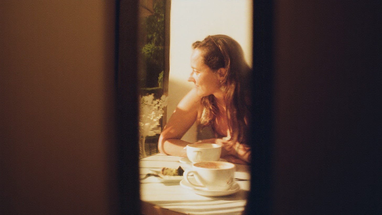 A woman with long hair sits at a café table in warm light, looking out of frame with two cups of coffee and a plate of dessert in front of her. Photo by Georges Yazbek