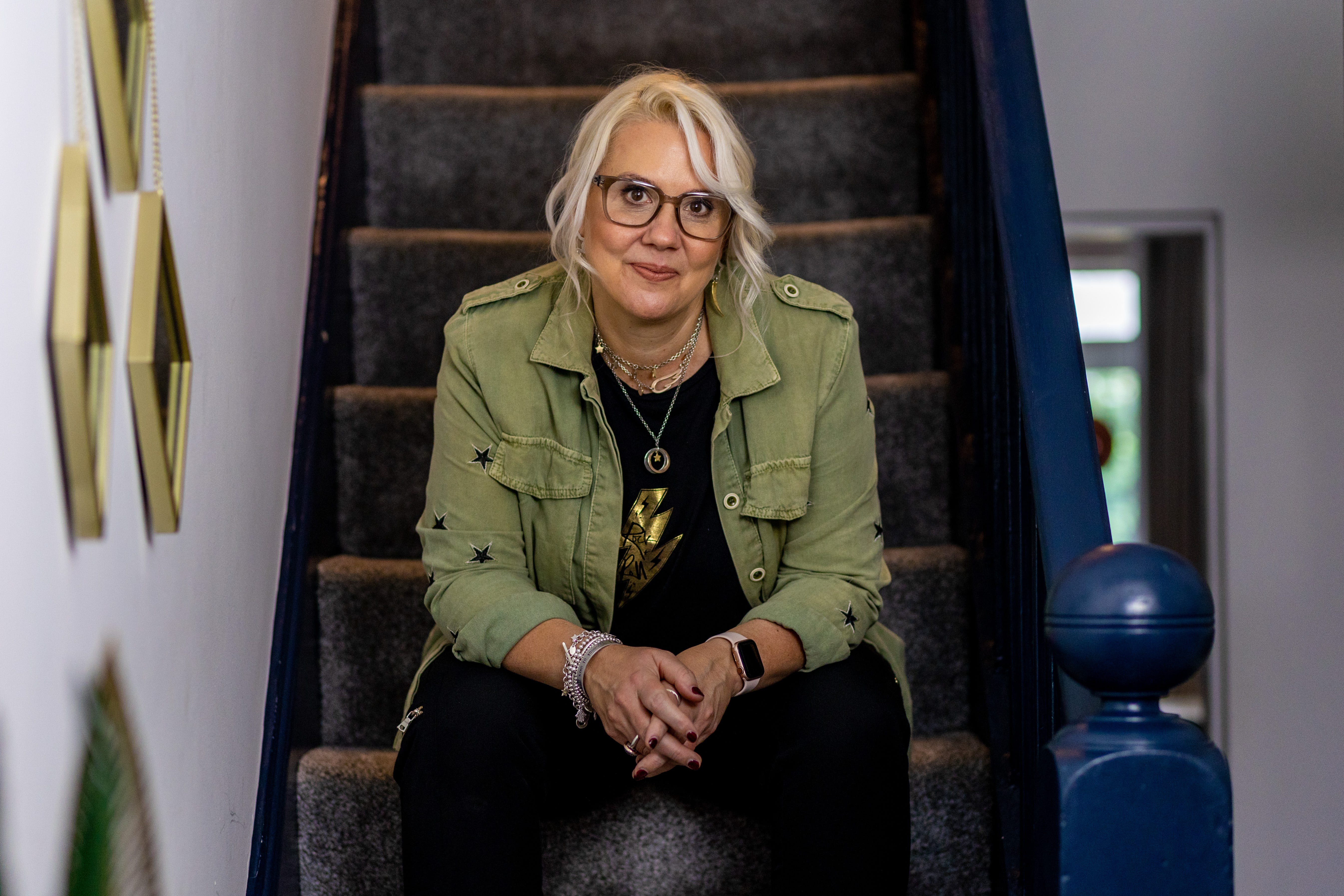 Sarah Knight is sat on her stairs. She has blonde wavy hair, large framed glasses and is wearing all-black plus a khaki jacket. 