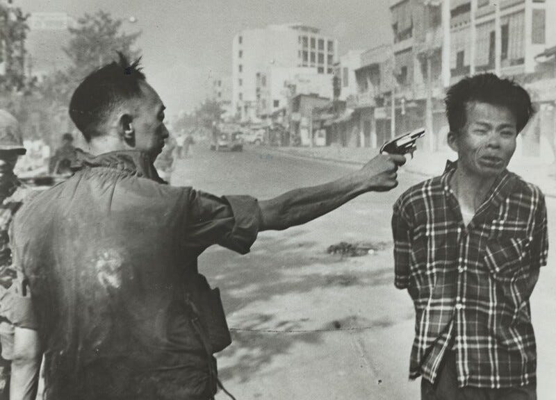 This is a black-and-white photograph of two men on a street lined with buildings. Both men are seen from the waist up. The man on the left has his back to us and looks to the left. He wears a shirt or jacket with a high collar and rolled up sleeves, and his hair is short. His arms are outstretched, pointing a small gun towards the man on the right. The man on the right faces us, with his hands behind his back. His face is contorted in an expression of distress, with his downturned mouth partially open. He wears a loose flannel shirt, and his hair is long on the top and shorter on the sides. both men have black hair and medium-toned skin. The street stretches beyond the men, towards the left of center, and it is lined with indistinct buildings in the top right corner of the photograph. On the left side, some trees can be seen, and a man in a helmet is partially visible.