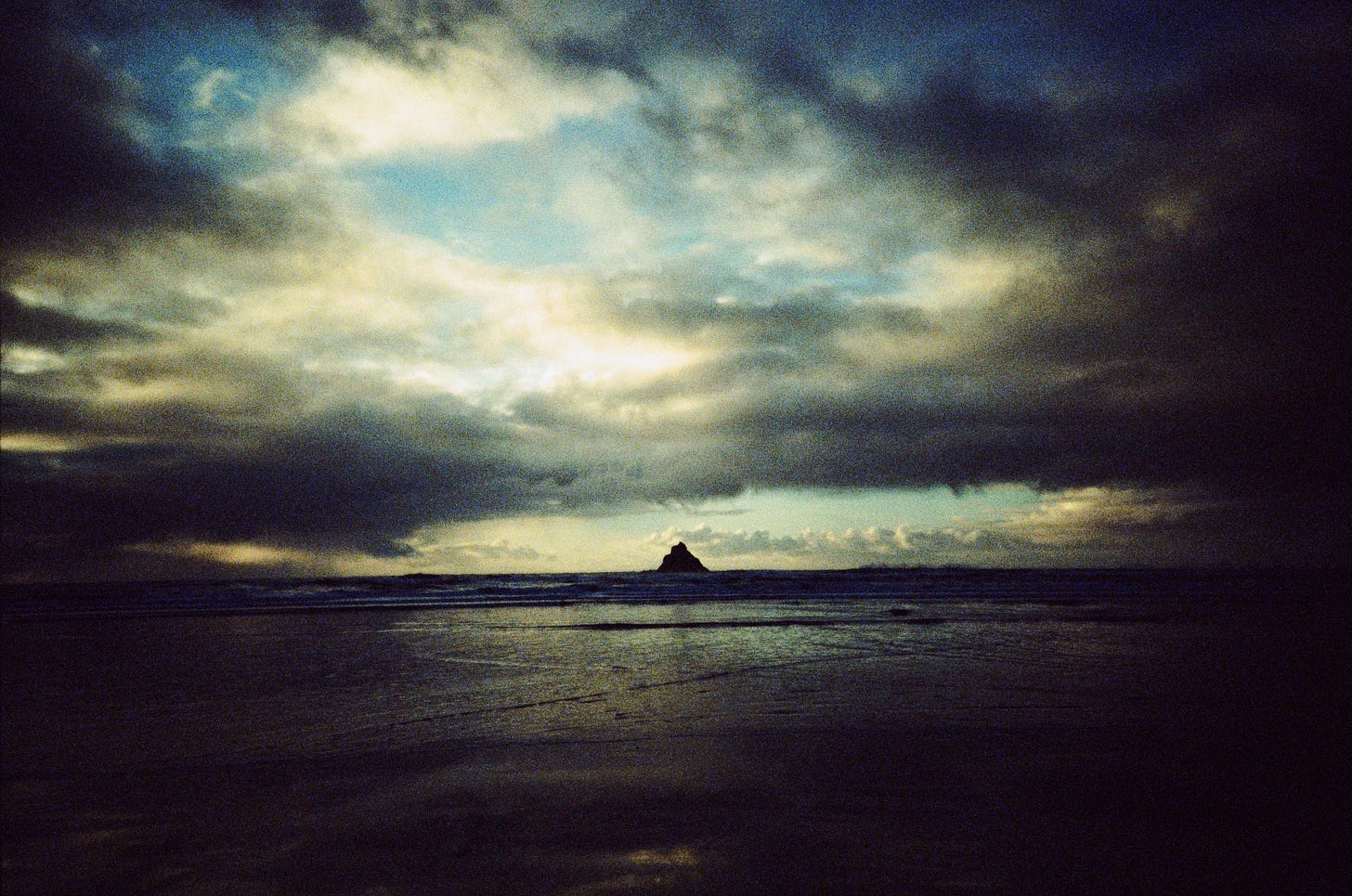 A dark & grainy film photo of the ocean, with a cloudy and colorful sky in the background. In the water in the far distance, a large triangular rock formation can be seen.